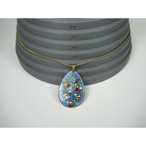 Puede incluir: Un collar con colgante en forma de l&aacute;grima con un dise&ntilde;o de esmalte azul con acentos coloridos. El colgante cuelga de una cadena dorada. El collar se muestra sobre una superficie gris escalonada con medidas en pulgadas.