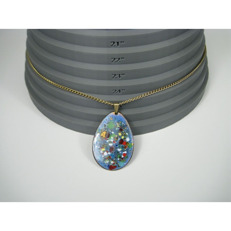 Puede incluir: Un collar con colgante en forma de l&aacute;grima con un dise&ntilde;o de esmalte azul con acentos coloridos. El colgante cuelga de una cadena dorada. El collar se muestra sobre una superficie gris escalonada con medidas en pulgadas.