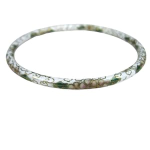 May include: A thin, circular bangle bracelet featuring a white enamel base decorated with a repeating pattern of small pink flowers, green leaves, and gold-toned outlines. The jewelry piece has a smooth, polished finish and a consistent, slender profile.