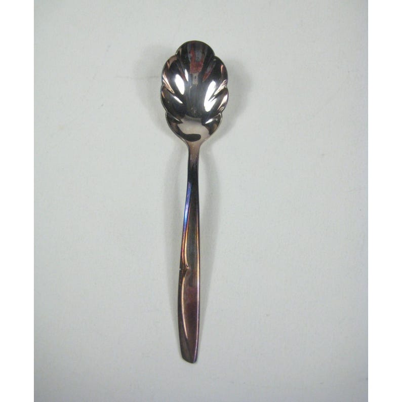 May include: A silver-colored spoon with a decorative, scalloped bowl. The spoon's handle is slender and tapers towards the end. The spoon is designed for serving or eating. The spoon is made of a shiny, reflective metal.