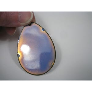 Puede incluir: Un colgante en forma de l&aacute;grima con un borde de bronce met&aacute;lico y una superficie iridiscente azul y dorada. El colgante est&aacute; sujeto por una mano, mostrando su color y dise&ntilde;o &uacute;nicos. El colgante tiene un peque&ntilde;o agujero en la parte superior para una cadena.