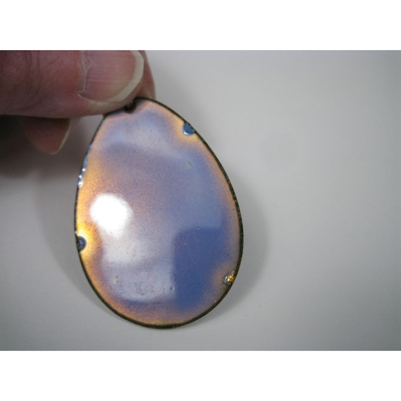 Puede incluir: Un colgante en forma de l&aacute;grima con un borde de bronce met&aacute;lico y una superficie iridiscente azul y dorada. El colgante est&aacute; sujeto por una mano, mostrando su color y dise&ntilde;o &uacute;nicos. El colgante tiene un peque&ntilde;o agujero en la parte superior para una cadena.