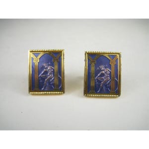 May include: A pair of rectangular cufflinks featuring a gold-toned frame with a beaded edge. The center displays a deep blue enamel background with a raised metallic figure seated between two gold-toned columns under a gothic arch design.