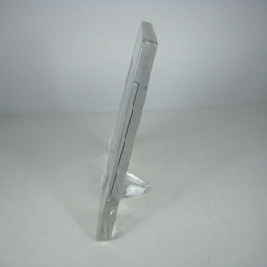 May include: A rectangular, clear glass picture frame standing upright in a side profile view. The frame features a smooth, transparent surface with a sturdy, angled base support that allows it to remain in a vertical position.