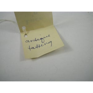 Pu&ograve; includere: Primo piano di un'etichetta gialla con le parole scritte a mano "antique fastening" in inchiostro blu. L'etichetta &egrave; attaccata a un filo bianco e si trova su uno sfondo bianco.