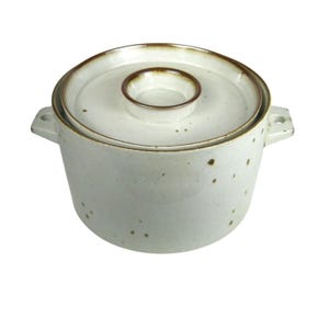 May include: A round ceramic casserole dish with a matching lid, featuring a speckled off-white glaze and brown rim accents. The pot includes two side handles and a central knob on the lid, showcasing a simple, functional design with earthy tones.
