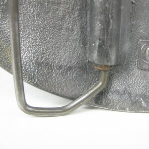 K&ouml;nnte beinhalten: Eine silberne G&uuml;rtelschnalle aus Metall mit einer strukturierten Oberfl&auml;che und einer Metallschlaufe. Die Schnalle hat einen kleinen, runden, kupferfarbenen Stift.