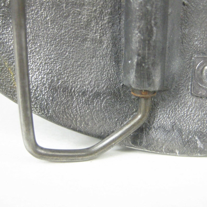 K&ouml;nnte beinhalten: Eine silberne G&uuml;rtelschnalle aus Metall mit einer strukturierten Oberfl&auml;che und einer Metallschlaufe. Die Schnalle hat einen kleinen, runden, kupferfarbenen Stift.