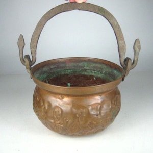 May include: A copper cauldron with a decorative embossed design around the body. The cauldron has a metal handle and is likely used for cooking or decorative purposes. The interior shows signs of age and use.