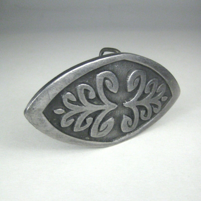 K&ouml;nnte beinhalten: Eine ovale G&uuml;rtelschnalle aus Silber mit einem komplizierten, gepr&auml;gten Design eines stilisierten Blattes.