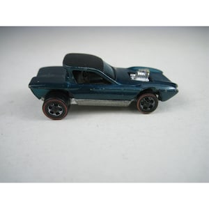 May include: A dark teal die-cast toy car featuring a black roof, a silver-colored engine block protruding from the hood, and four black wheels with thin red line accents along the outer edges of the tires, viewed from the side profile.