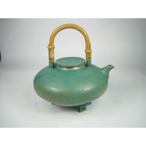 May include: A teal ceramic teapot with a rounded, low-profile body, a matching lid, and a short spout. The teapot features a curved, light-coloured bamboo handle attached to two upright ceramic loops on the top, and it stands on three small feet.