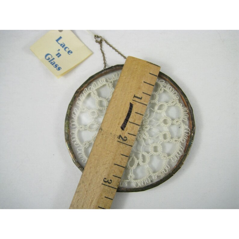 Pu&ograve; includere: Un centrino rotondo in pizzo color crema incorniciato in un anello di metallo argentato. Un righello di legno &egrave; posto sul centrino, misurando circa 7,6 cm. Un'etichetta recita "Lace 'n Glass".