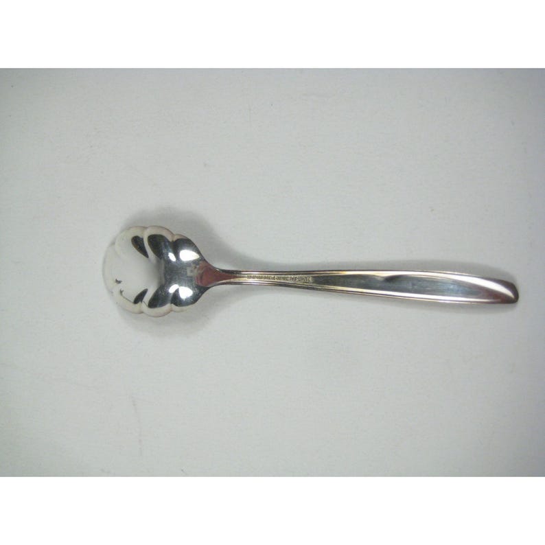 May include: A silver-colored spoon with a scalloped bowl and a long, slender handle. The spoon is made of a shiny, reflective metal, likely stainless steel or silver. The design is simple and elegant, suitable for serving sugar or other condiments.