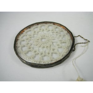 Pu&ograve; includere: Un acchiappasogni rotondo in stile vintage con un centro in pizzo all'uncinetto color crema. Il pizzo &egrave; incorniciato da vetro trasparente e un bordo in metallo scuro. Una piccola catena e un'etichetta sono attaccate al telaio.