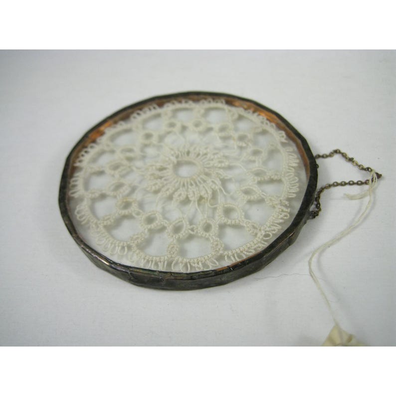 Pu&ograve; includere: Un acchiappasogni rotondo in stile vintage con un centro in pizzo all'uncinetto color crema. Il pizzo &egrave; incorniciato da vetro trasparente e un bordo in metallo scuro. Una piccola catena e un'etichetta sono attaccate al telaio.