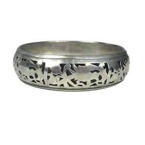 May include: A silver-toned metal bangle bracelet featuring a continuous cut-out pattern of stylized floral and leaf motifs. The bracelet has a smooth, polished upper and lower rim, with a dark, recessed background behind the intricate metalwork.