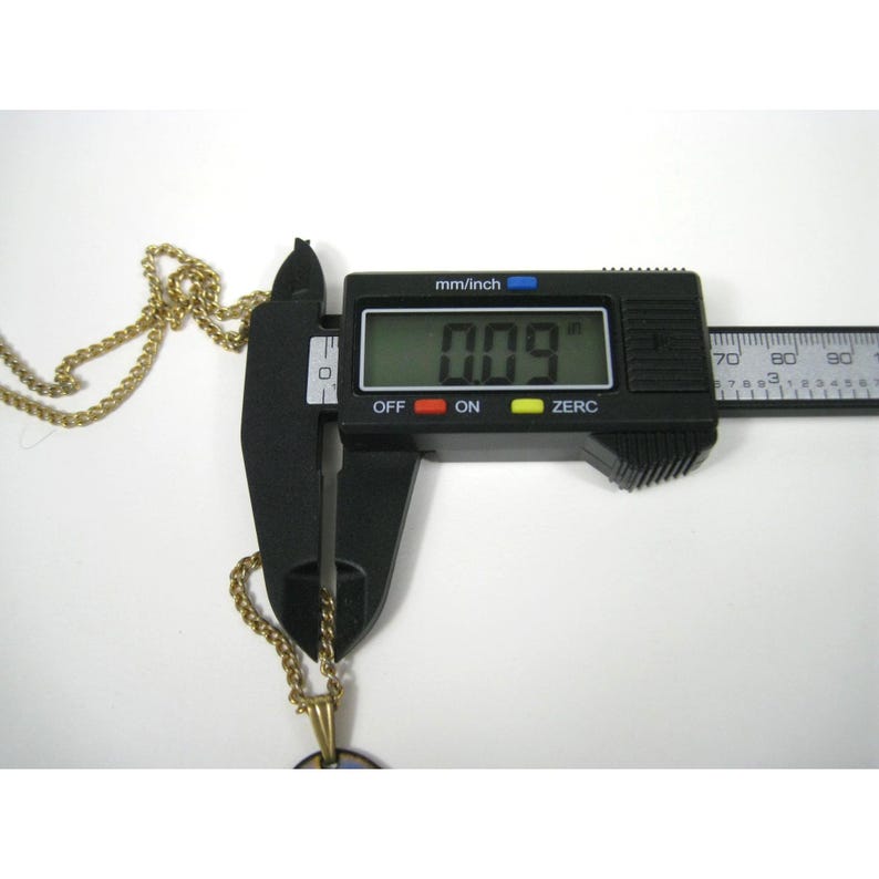 Puede incluir: Un collar dorado con un colgante se mide con un calibrador digital negro. El calibrador muestra una medida de 2,29 mm. La cadena del collar es visible en el lado izquierdo de la imagen.