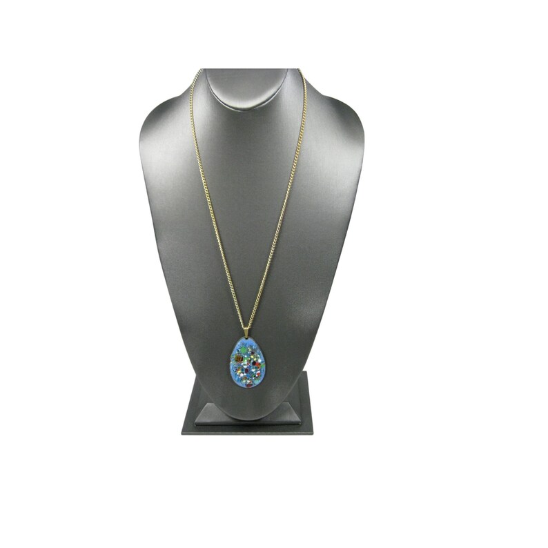 Puede incluir: Un collar de cadena dorada con un colgante azul. El colgante tiene forma de l&aacute;grima y presenta un dise&ntilde;o de mosaico con piedras preciosas de colores. El collar se exhibe en un busto de joyer&iacute;a gris.