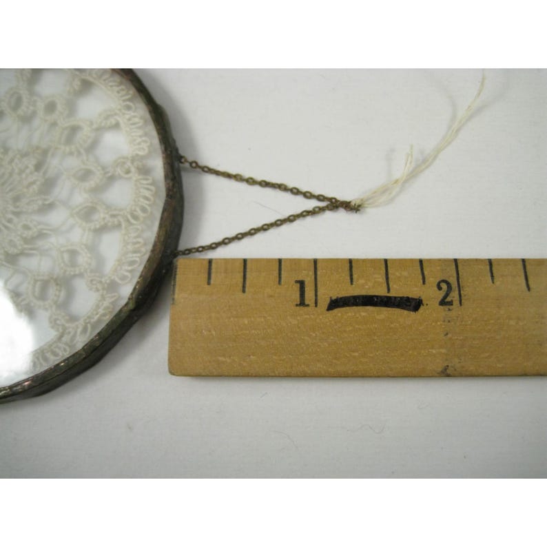 Pu&ograve; includere: Un oggetto rotondo in vetro trasparente con un motivo in pizzo color crema. L'oggetto &egrave; attaccato a una piccola catena di metallo. Un righello di legno &egrave; posto accanto. Il righello misura in centimetri.