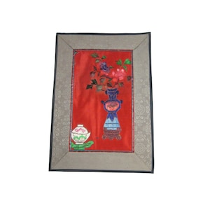 Puede incluir: Una obra de arte bordada enmarcada que presenta un jarrón de flores sobre un fondo rojo. El jarrón es azul y blanco con un diseño floral. Las flores son rosas, rojas y blancas. El marco es gris con un patrón floral.