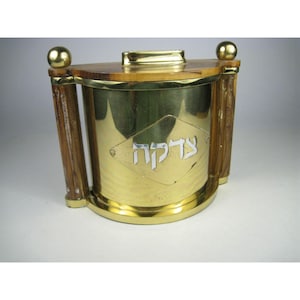 Puede incluir: Una caja de tzedakah semicircular construida en latón pulido y madera. El frente presenta una placa en forma de diamante con la palabra hebrea Tzedakah grabada en blanco. Incluye dos pilares verticales de madera y una ranura superior.