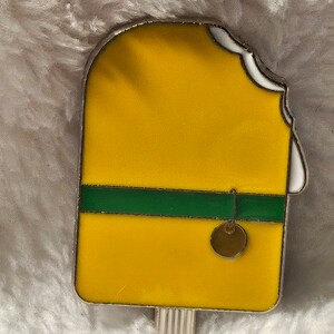 May include: A yellow popsicle with a green stripe and a small gold charm hanging from it. The popsicle is partially eaten, with a bite taken out of the top.