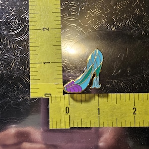 May include: A decorative enamel pin shaped like a high-heeled shoe. The pin is about 2.5 cm long and has a gold outline with blue, green, and purple details. The shoe design is reminiscent of a fairy tale.