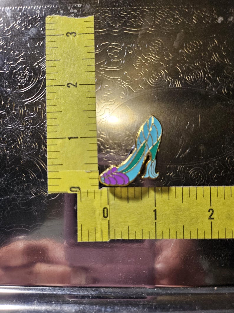 May include: A decorative enamel pin shaped like a high-heeled shoe. The pin is about 2.5 cm long and has a gold outline with blue, green, and purple details. The shoe design is reminiscent of a fairy tale.