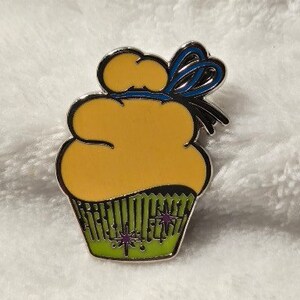 May include: A yellow cupcake with a blue bow and green frosting with purple stars. The cupcake is a pin.