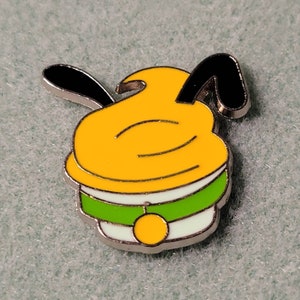 May include: A yellow and green enamel pin featuring a cartoon dog with floppy ears. The dog is wearing a green collar with a yellow tag.