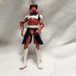 May include: A red and white action figure of a soldier in a helmet and armor. The figure has black accents on the waist and shoulders, and white boots. The figure is standing with its arms at its sides.