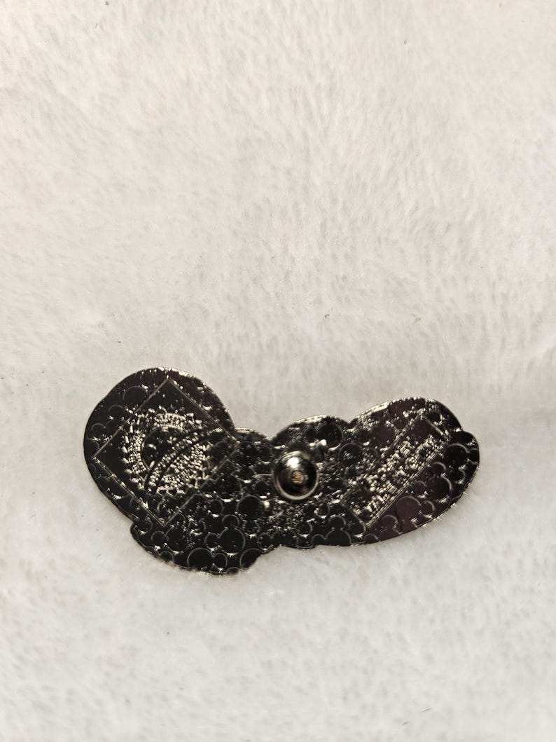 Puede incluir: Un pin de metal gris oscuro con un dise&ntilde;o de mariposa. El pin tiene una superficie texturizada y un respaldo de alfiler para la fijaci&oacute;n. El dise&ntilde;o de la mariposa es detallado e intrincado, con patrones visibles en las alas.