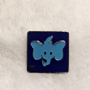 May include: A square enamel pin featuring a light blue elephant design on a dark blue background. The elephant has large ears and a trunk. A small black paw print is in the lower left corner. The pin is likely a collectible item.