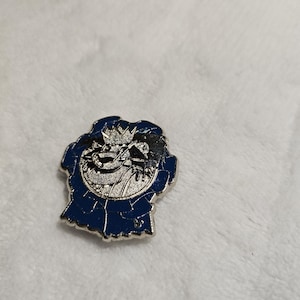 May include: A silver-colored pin with a blue border. The pin features a detailed illustration of a dragon wearing a crown. The dragon is set against a circular background with intricate detailing.