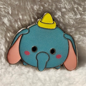 May include: A blue and pink Dumbo pin with a yellow hat. The pin is a cartoon version of the Disney character Dumbo.