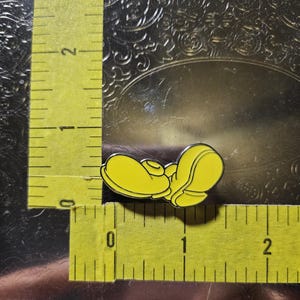 Puede incluir: Un pin de esmalte amarillo con forma de zapato de dibujos animados. El pin mide aproximadamente 2,5 cm de largo y est&aacute; sobre un fondo met&aacute;lico texturizado. El pin tiene un contorno negro.