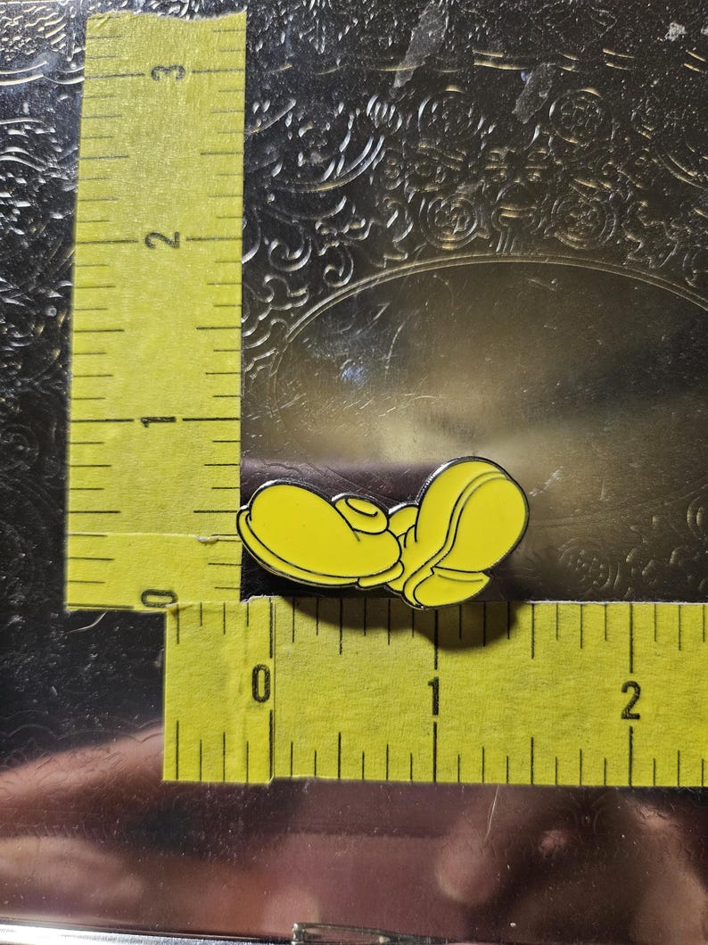 Puede incluir: Un pin de esmalte amarillo con forma de zapato de dibujos animados. El pin mide aproximadamente 2,5 cm de largo y est&aacute; sobre un fondo met&aacute;lico texturizado. El pin tiene un contorno negro.