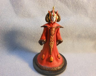 May include: A figurine of a character wearing a long red gown with gold embroidery, a high collar, and a headpiece with dark brown curved side elements. The gown features a flared skirt with orange oval accents and a dark brown base stand.