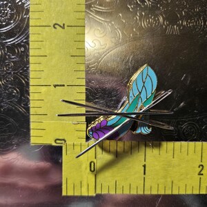 May include: A butterfly-shaped enamel pin with a teal and purple design, accented with gold-coloured metal. The pin measures approximately 2.5 cm in length and is set against a textured, metallic background.
