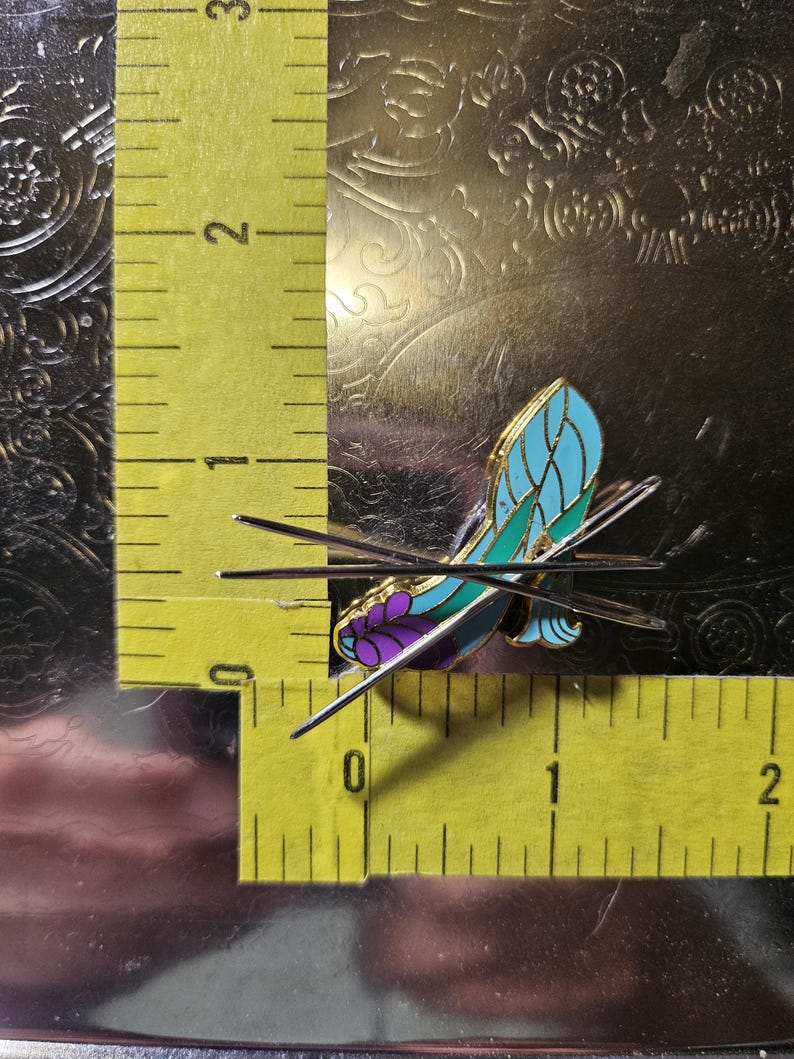 May include: A butterfly-shaped enamel pin with a teal and purple design, accented with gold-coloured metal. The pin measures approximately 2.5 cm in length and is set against a textured, metallic background.