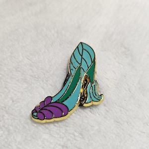 May include: A decorative enamel pin shaped like a high-heeled shoe. The pin has a gold border and is filled with teal, green, and purple enamel. It's a fashion accessory or a collectible item.