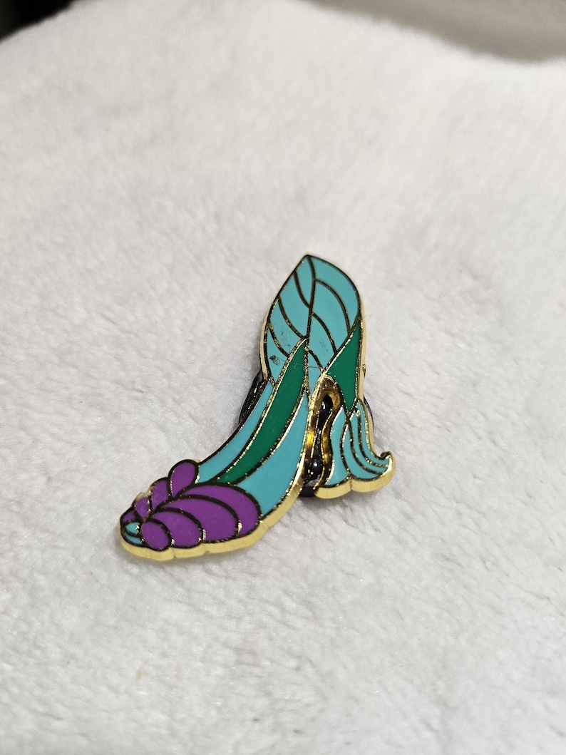 May include: A decorative enamel pin shaped like a high-heeled shoe. The pin has a gold border and is filled with teal, green, and purple enamel. It's a fashion accessory or a collectible item.