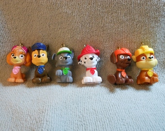 May include: A collection of six Paw Patrol character figurines. Each figurine is a different color and features a unique hat or helmet. The figurines are made of plastic and have a gold loop at the top for hanging.