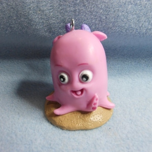 May include: A pink octopus figurine with large white eyes, black pupils, and a small open mouth, featuring purple accents on top and a metal loop for hanging. The creature is positioned on a textured, tan, circular base with a sandy appearance.