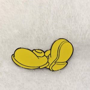 Puede incluir: Un pin de esmalte amarillo que representa un pie de dibujos animados estilizado, con un contorno negro. El pin es de color amarillo brillante y est&aacute; sobre un fondo blanco texturizado. Es probable que el pin sea un art&iacute;culo de colecci&oacute;n o un accesorio.