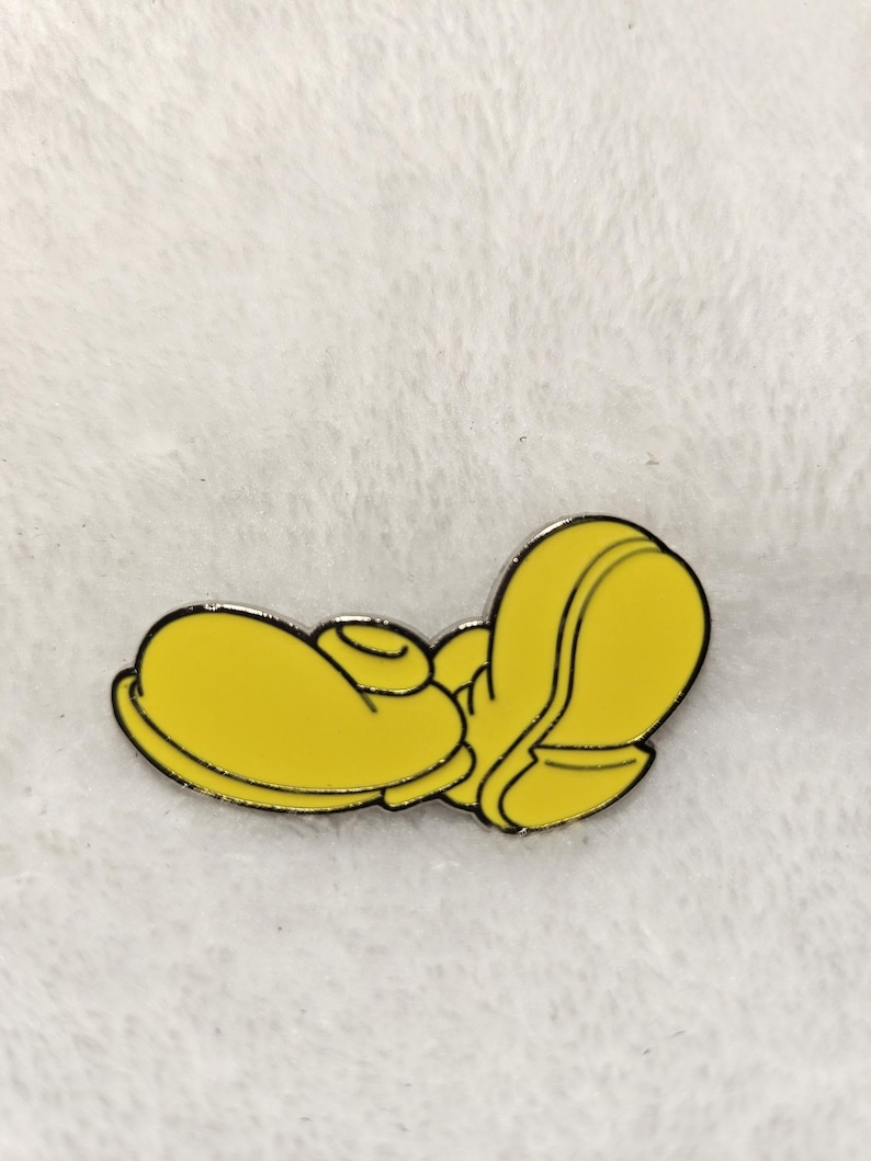 Puede incluir: Un pin de esmalte amarillo que representa un pie de dibujos animados estilizado, con un contorno negro. El pin es de color amarillo brillante y est&aacute; sobre un fondo blanco texturizado. Es probable que el pin sea un art&iacute;culo de colecci&oacute;n o un accesorio.