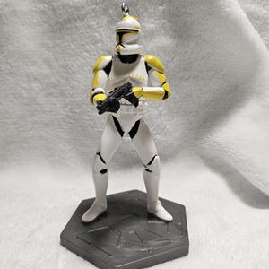 May include: A white and yellow figurine of a clone trooper from Star Wars, holding a black blaster. The figure is standing on a gray hexagonal base. The trooper's helmet and armor have yellow accents.