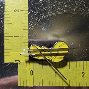 Puede incluir: Un pin de esmalte amarillo y negro con forma de abeja estilizada, colocado sobre una regla amarilla. El pin mide aproximadamente 2 cm de largo. La regla, marcada en pulgadas, est&aacute; sobre una superficie reflectante.