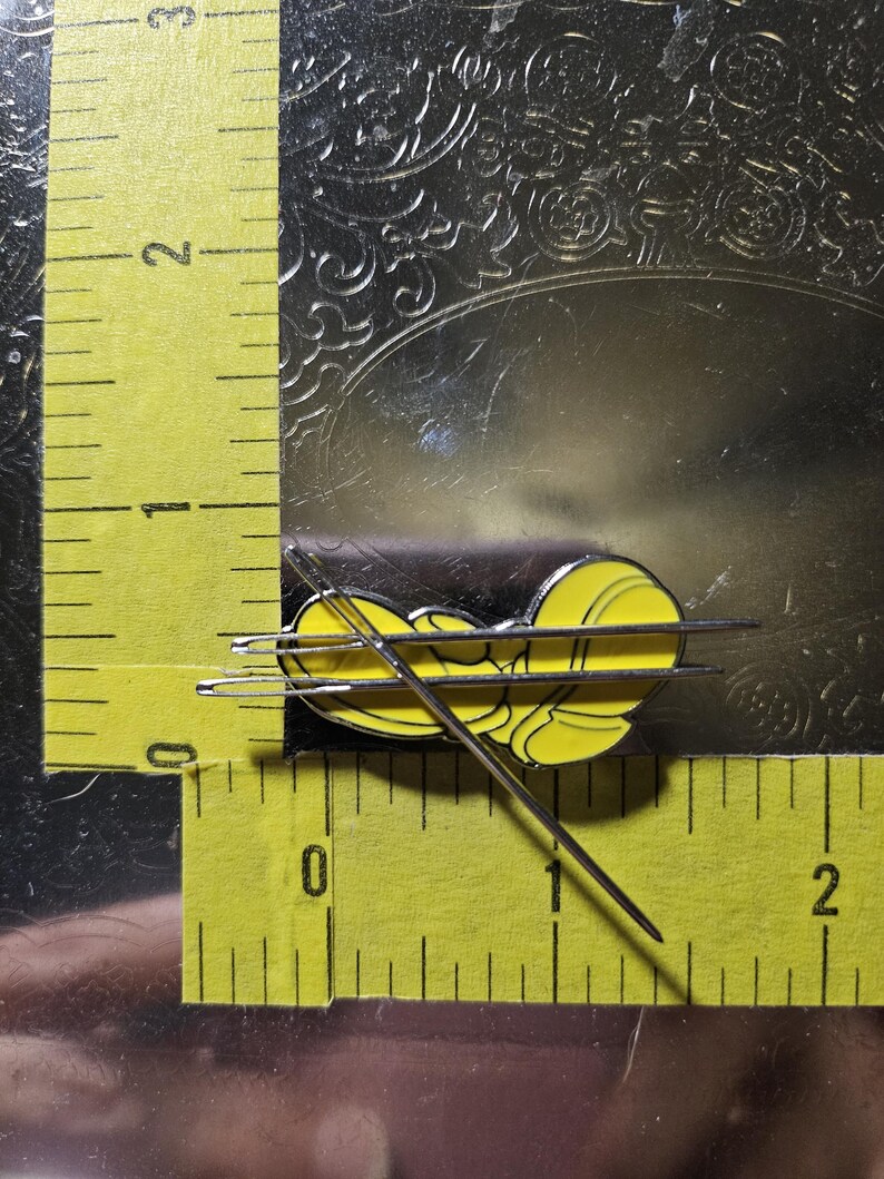 Puede incluir: Un pin de esmalte amarillo y negro con forma de abeja estilizada, colocado sobre una regla amarilla. El pin mide aproximadamente 2 cm de largo. La regla, marcada en pulgadas, est&aacute; sobre una superficie reflectante.